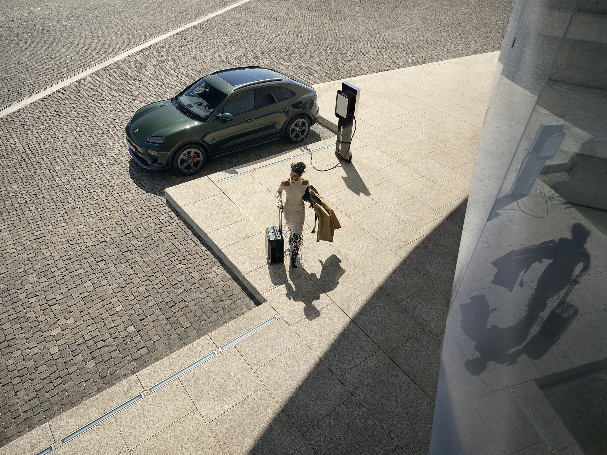 Green Porsche electric SUV charging at modern EV station while traveler with suitcase walks by. Premium electric mobility, luxury design, and sustainable charging solution for business and leisure travel.