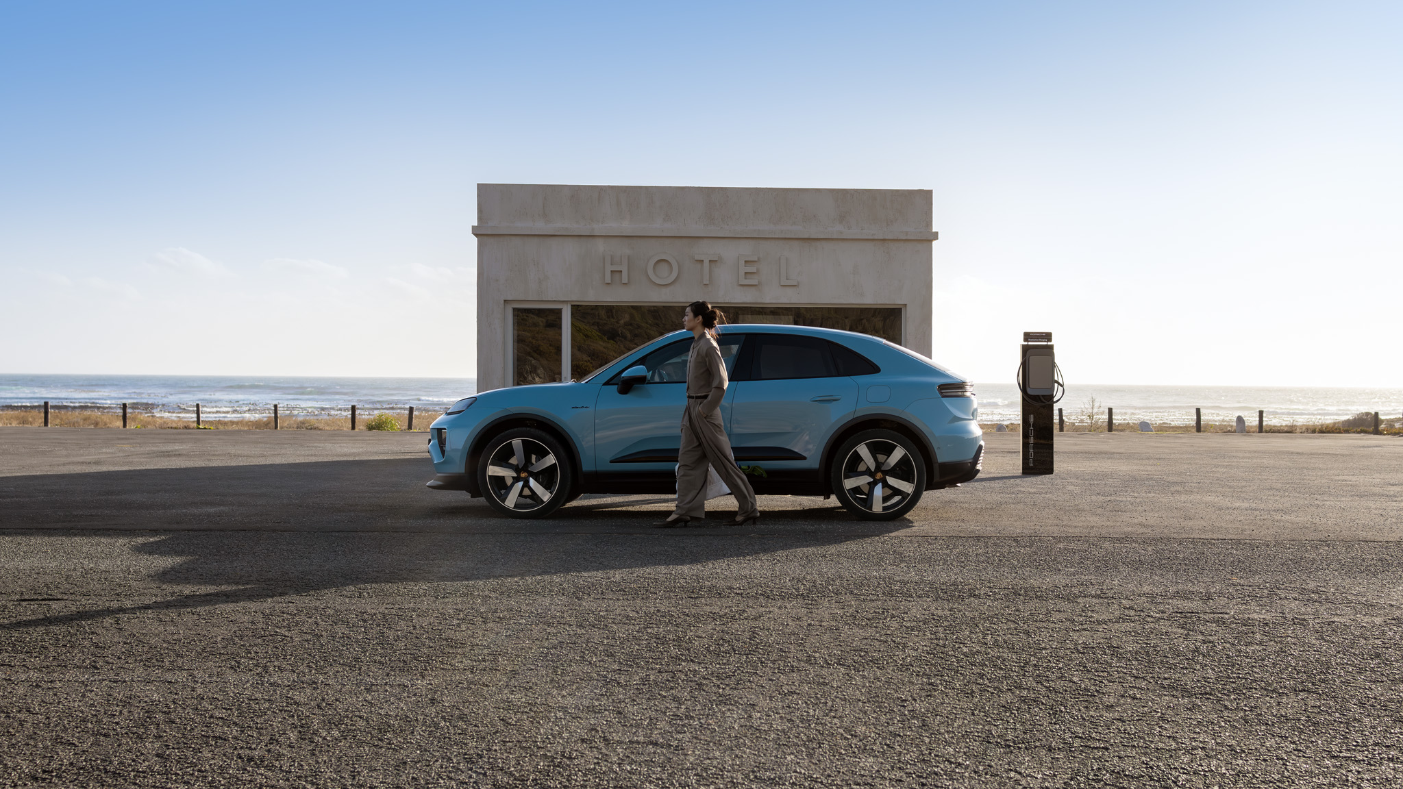 Eine Frau geht an ihrem blauen Porsche Macan Electric vorbei, an dessen Seite eine Ladestation zu sehen ist.