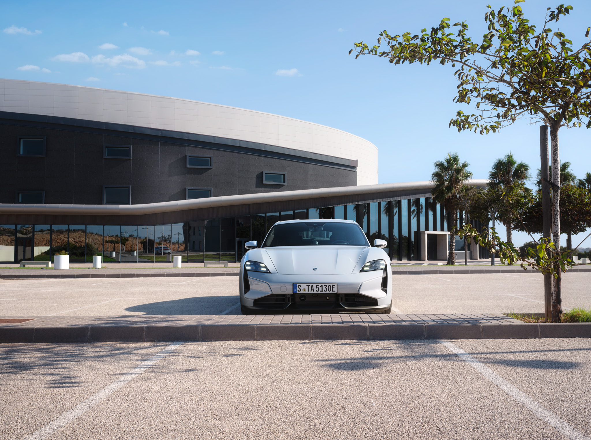 Vue de face d’une Porsche blanche garée dans une place de parking vide devant un bâtiment moderne à l’architecture courbe et aux grandes fenêtres vitrées, entourée de palmiers sous un ciel bleu clair.
