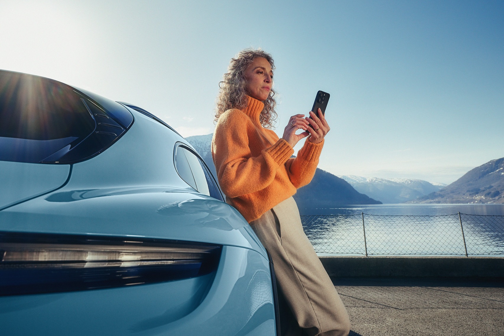 Vrouw in een oranje trui die tegen een blauwe Porsche leunt terwijl ze een smartphone gebruikt, met een meer en bergen op de achtergrond.