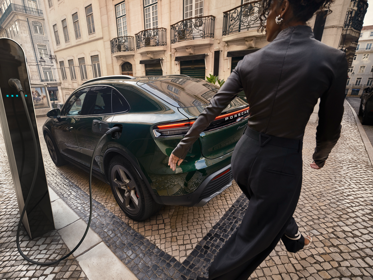 Ein Macan Electric ist in der Stadt an einer Ladestation angeschlossen, während eine Person auf das Fahrzeug zugeht.