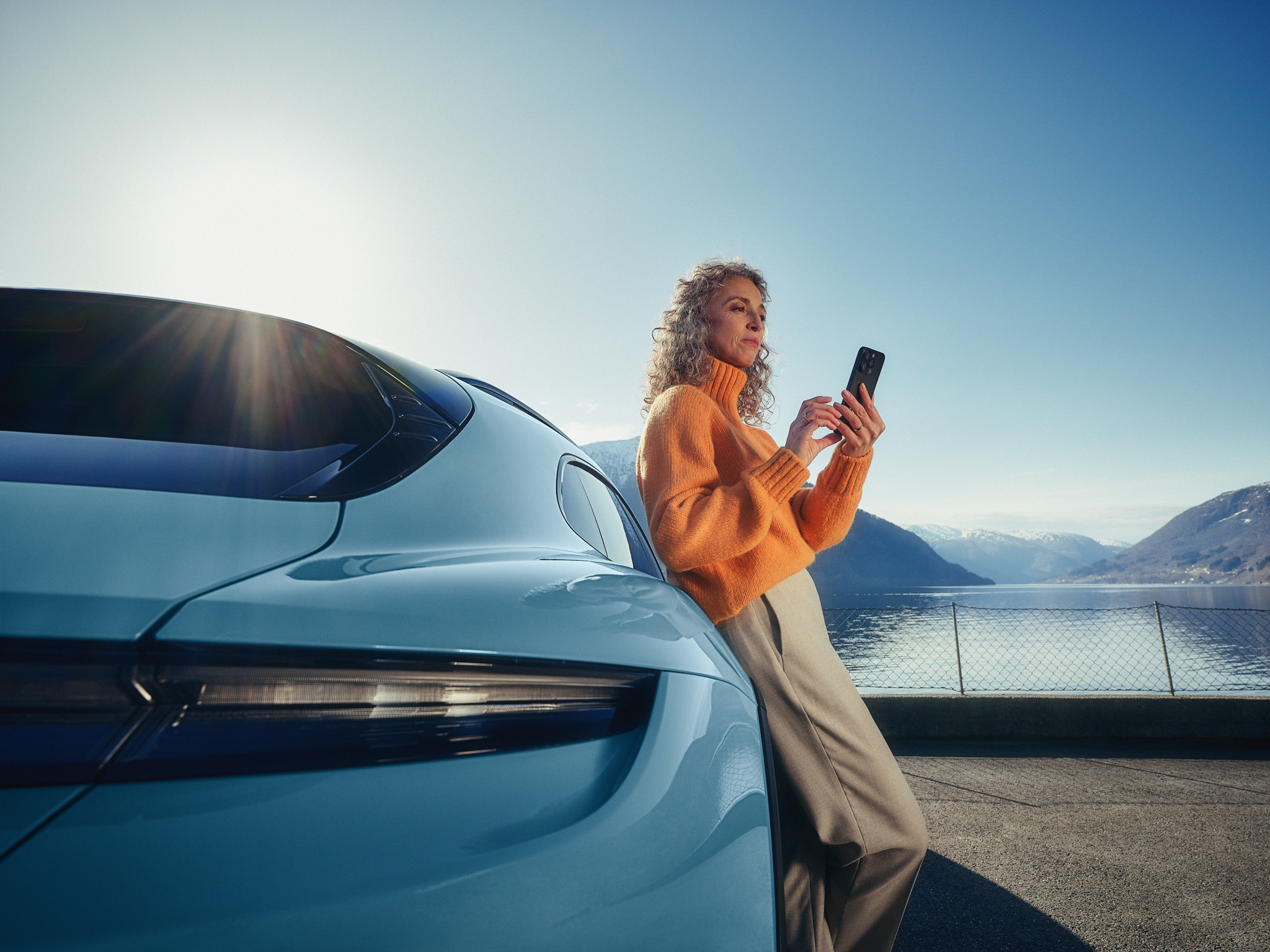 A woman with a smartphone in her hand leans against a light blue Porsche Macan Electric.