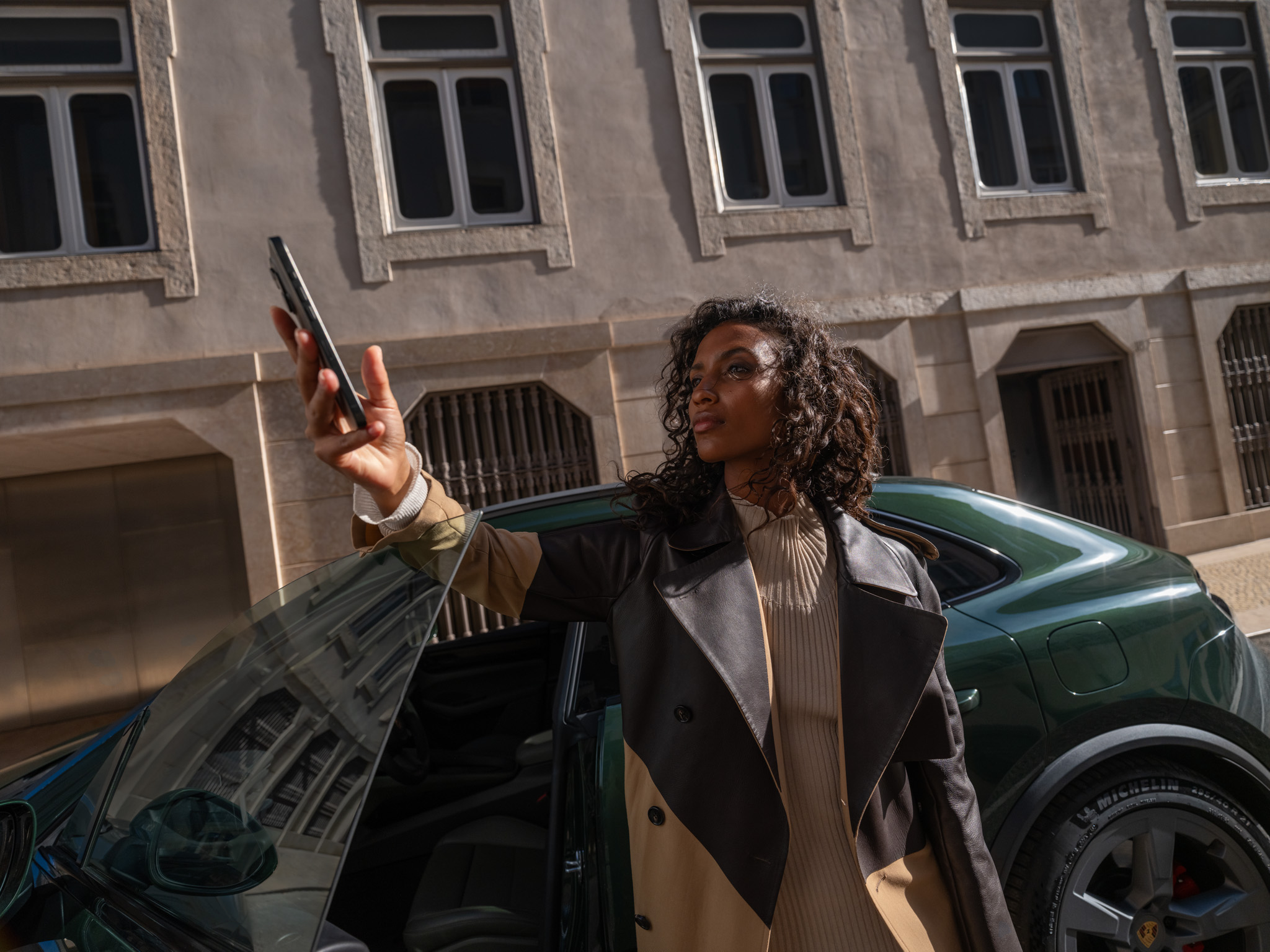 Eine Frau steht mit einem Smartphone in der Hand vor einem grünen Macan Electric mit geöffneter Fahrertür.