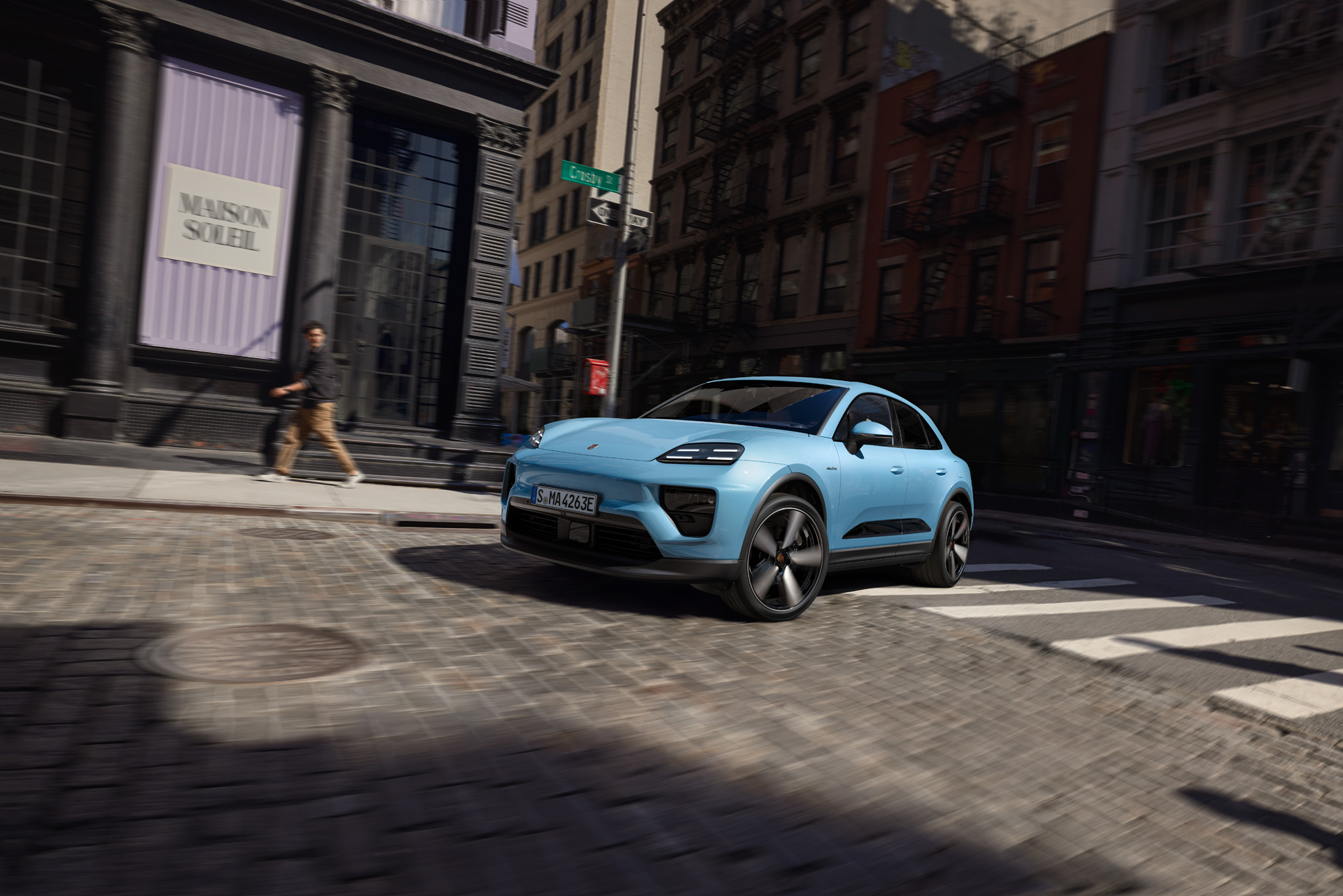 Ein blauer Macan Electric fährt über einen Zebrastreifen in einer Kurve, und eine Person dreht sich um und schaut auf das Fahrzeug.